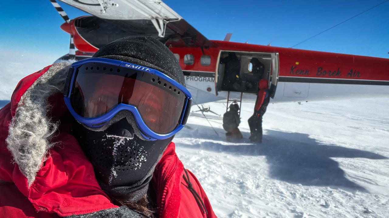 A woman with goggles standing on an ice sheet with a plane behind her.
